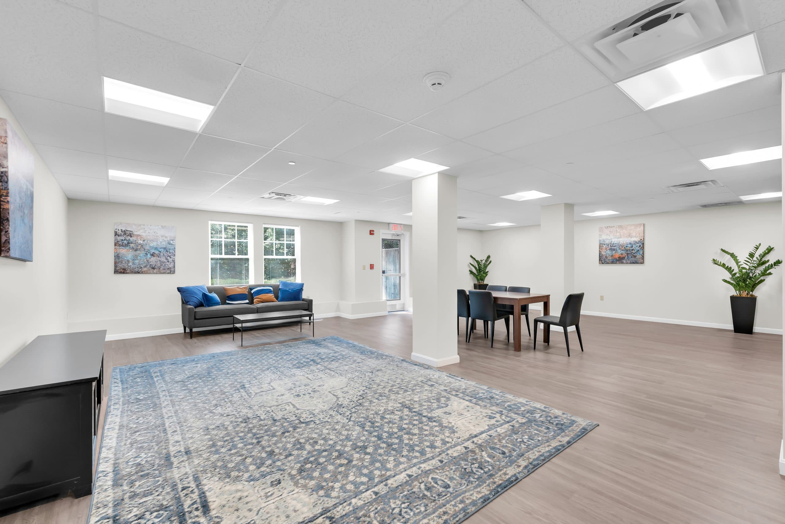 The Barton Commons community room — a bright open space with a sofa, a dining table for six, and a patterned rug over plank flooring