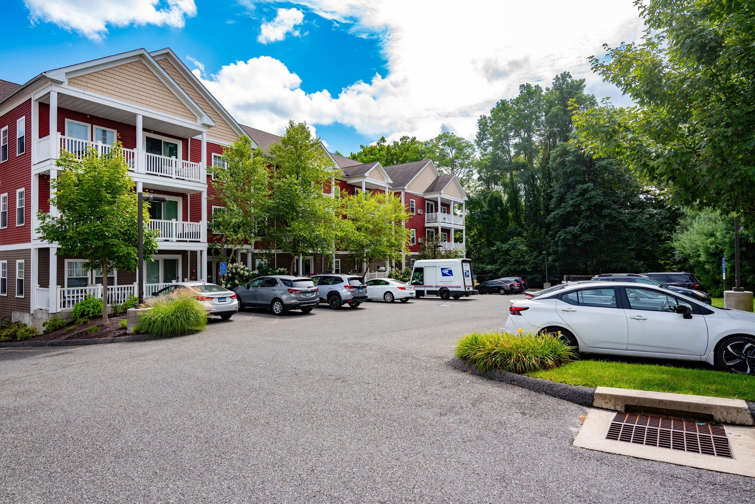 Barton Commons on a summer afternoon — red and tan clapboard apartment buildings with white porch balconies, seen across the resident parking court